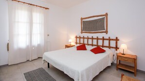 A bedroom with a bed, two bedside tables with lamps, a wooden headboard, a patterned wall hanging, and a window with sheer curtains. - Vacation Home "Trilo Mare" with Sea View, Private Terrace & Wi-Fi (Budoni)