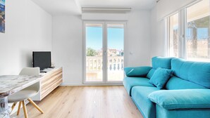 Una sala moderna con piso de madera, un sofá azul, un televisor sobre un mueble de madera y un balcón con vista a edificios.