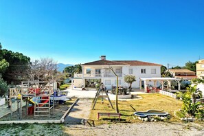 Un bâtiment blanc avec un toit en tuiles, une aire de jeux avec des balançoires et une petite piscine.