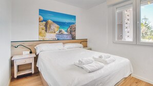Un dormitorio con cama, ropa de cama blanca, una mesita de noche, un cuadro colgado en la pared que representa una escena costera y una ventana con vistas a zonas verdes.