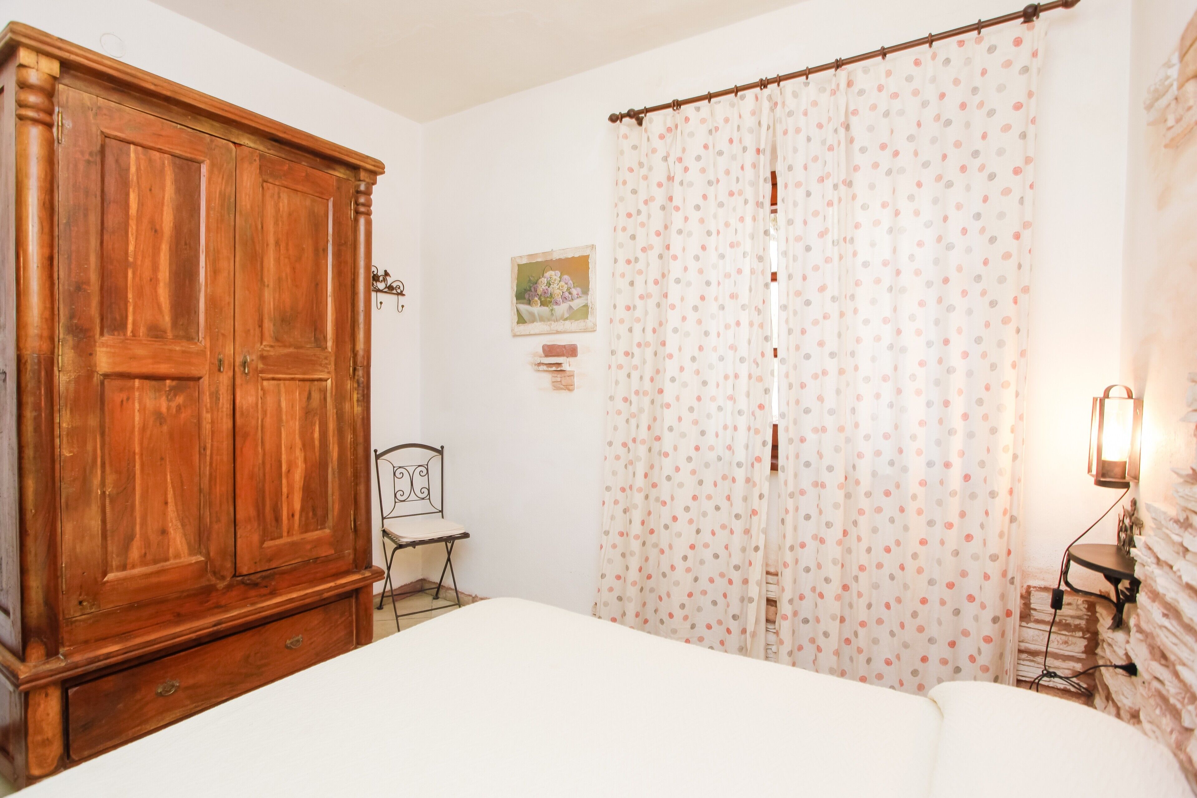 A bedroom with a wooden wardrobe, a bed, a chair, a polka dot curtain, and a wall-mounted lamp.