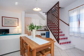 Un coin repas avec une table et des chaises en bois, un vase de fleurs, un téléviseur et, au fond, une cuisine.