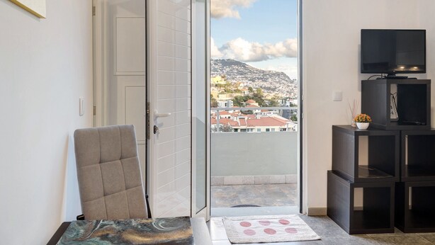 Een kamer met een schuiframen naar een balkon, een stoel, een klein tafeltje, een tv op een standaard en uitzicht op een stadsbeeld.