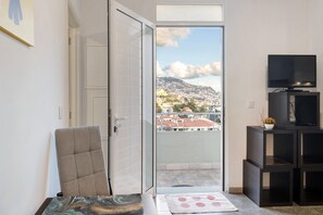 Una habitación con una puerta corrediza de vidrio que da a un balcón, una silla, una mesita, un televisor sobre un mueble bajo y vistas a un paisaje urbano.