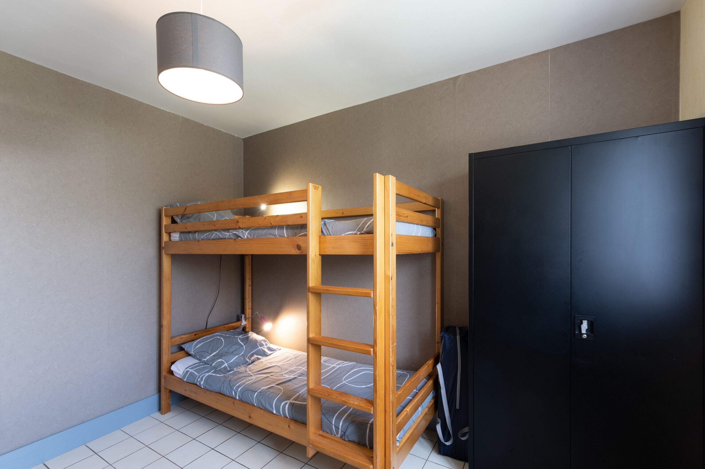 A bunk bed with a headboard, a bedside table, and a lamp. There is a wardrobe with a handle and a door.