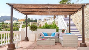 Een overdekt terras met rieten meubilair, een witte balustrade en een stenen trap.