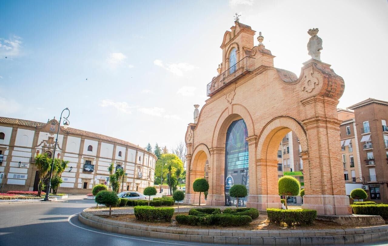 Un edificio histórico con entradas abovedadas y una torre del reloj, rodeado de un jardín paisajístico con arbustos podados.