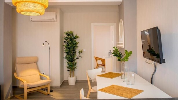 Una sala de estar moderna con un sofá, una mesa de comedor con sillas, una planta en maceta, un televisor montado en la pared y una lámpara colgante.