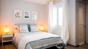 Un dormitorio con cama, lámparas de noche, una ventana con cortinas y cuadros en la pared.