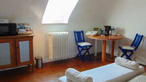 Una habitación pequeña y acogedora con una cama, dos sillas azules, una mesa con una cafetera, un radiador y un televisor sobre un mueble.