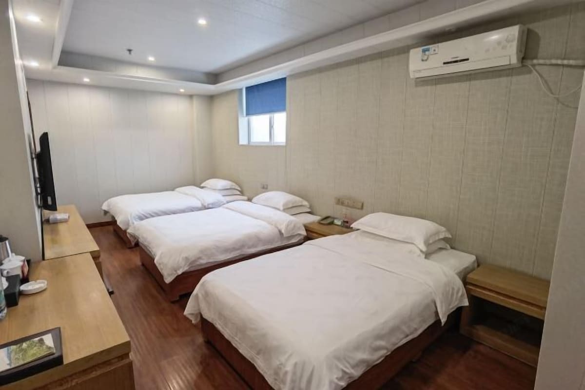 A hotel room with three single beds, a wooden desk, a flat-screen TV, and an air conditioning unit.