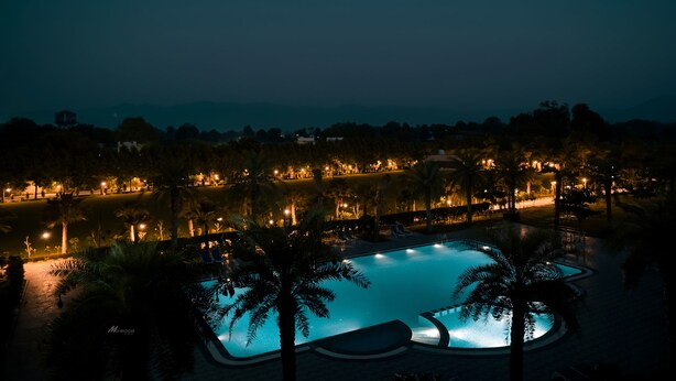 A nighttime view of a resort pool area with palm trees, a swimming pool, and surrounding buildings with lights. - Pride Resort Ranakpur. (Desuri)