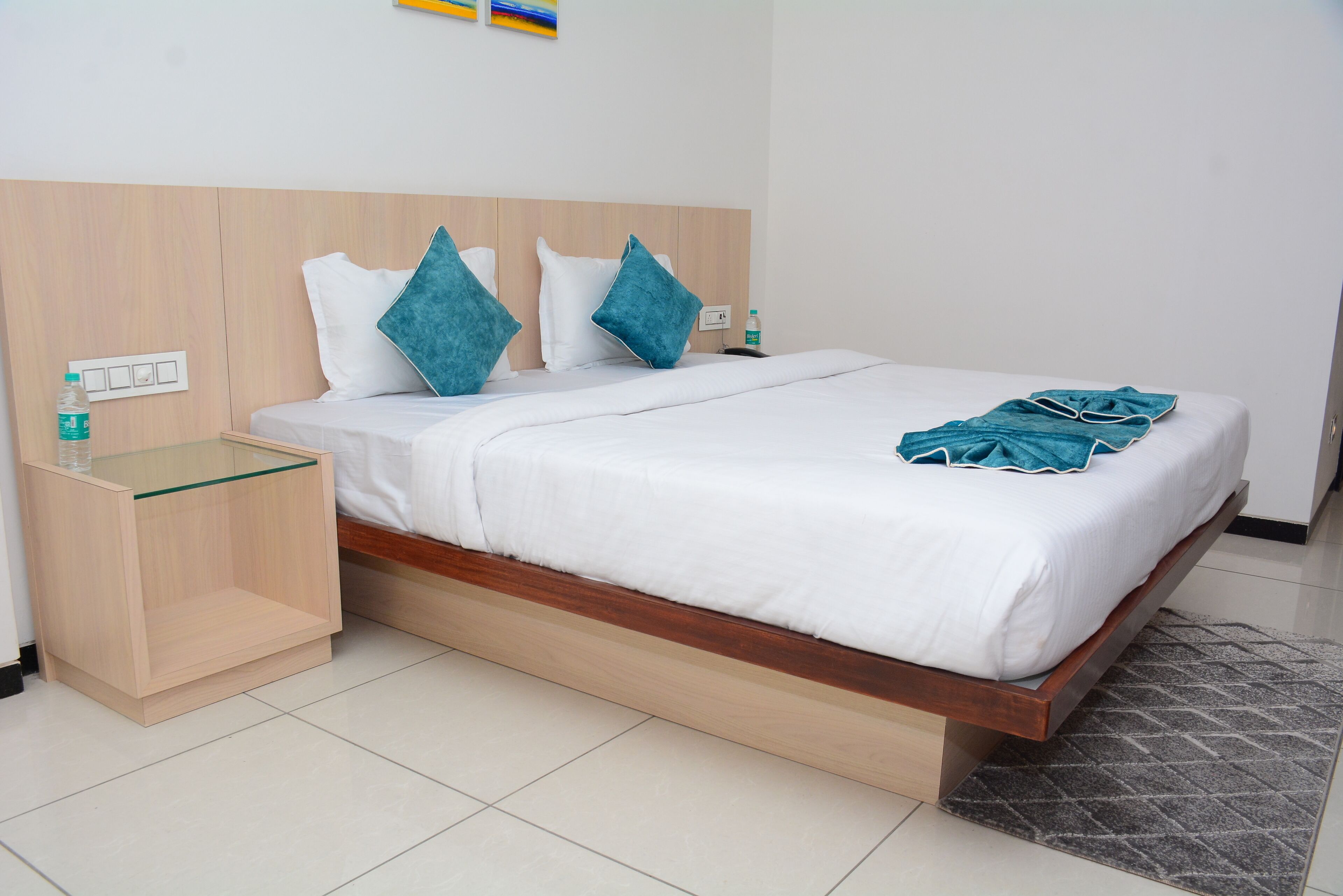 Une chambre d’hôtel avec une tête de lit en bois, une table de chevet en verre, un couvre-lit blanc, des coussins turquoise et un tapis gris à motifs.