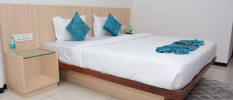 A hotel room with a wooden headboard, a glass bedside table, a white bedspread, teal pillows, and a grey patterned rug.