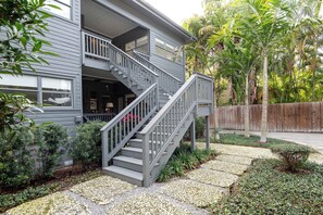 Una casa de dos pisos con fachada gris, una escalera de madera y un jardín con diversas plantas y cerca de madera.
