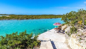 A coastal view with a clear blue sea, a concrete platform with a beach chair and umbrella, and lush greenery. - Private Pool & Stunning Sunsets Near Chalk Sound (Providenciales)