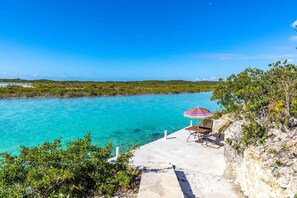 Une vue sur la côte, avec une mer bleue et limpide, une plateforme en béton sur laquelle se trouvent une chaise de plage et un parasol, ainsi qu’une végétation luxuriante.