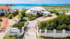 A large, white, two-story house with a red-tiled roof, surrounded by lush greenery and a view of the ocean. - Private Pool & Stunning Sunsets Near Chalk Sound (Providenciales)