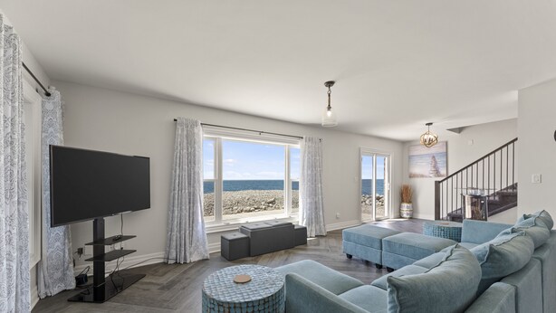 A modern living room with a large flat-screen TV, a blue sofa, and a view of the sea through large windows. - Cozy 3 Bedroom Lakefront Cottage (Blenheim)