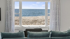 A room with a large window overlooking a rocky beach and the sea. There is a sofa with cushions in the foreground. - Cozy 3 Bedroom Lakefront Cottage (Blenheim)