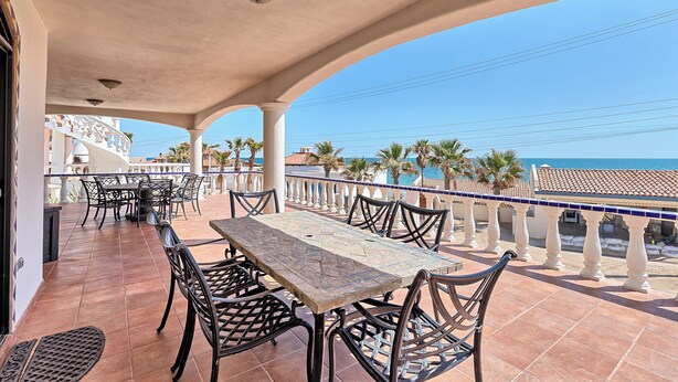 A covered outdoor terrace with a table and chairs, a tiled floor, and a view of the sea and buildings in the background.