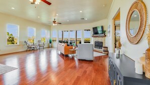 A spacious living room with hardwood floors, a ceiling fan, a fireplace, and large windows offering a view of the outdoors.