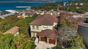 A two-story house with a stone foundation and a brown roof, surrounded by lush greenery and a view of the ocean.