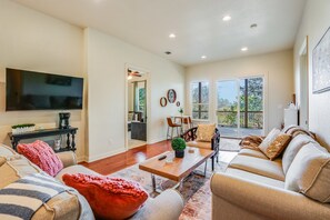 A spacious living room with a large flat-screen TV, a coffee table, and a sofa set. There is a dining area with chairs and a table, and a kitchen in the background.