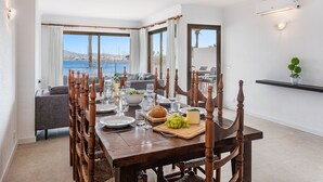 Un comedor con vistas al mar, una mesa de madera dispuesta con platos, vasos y una tabla de quesos.