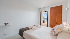Un dormitorio con cama, armario de madera, una pequeña estantería con una planta y vistas al exterior.