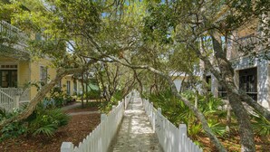 A pathway flanked by white picket fences and trees, leading to a building with a yellow facade and green shutters. - Seaside, FL, Steps to Pool! Walk to Beach, Sleeps 6 + 2 Adult Bikes! (Santa Rosa Beach Fl)