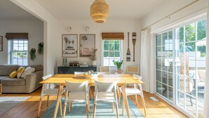 Amplia sala de estar con una mesa de comedor de madera, sillas blancas, un sofá y un ventanal con vistas al exterior.