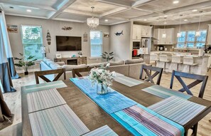A spacious living area with a large sofa, a dining table with placemats, a kitchen area, and a television mounted on the wall.