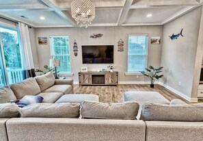 A living room with a sectional sofa, a flat-screen TV, decorative wall art, and a chandelier. - Tidal Walk * New 5BR Bethany Beach House (Millville)