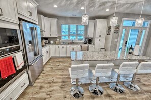 A modern kitchen with stainless steel appliances, white cabinetry, a large island with bar stools, and pendant lighting. - Tidal Walk * New 5BR Bethany Beach House (Millville)