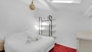 Een slaapkamer met dakraam, een bed met witte lakens, een roodgekleurd vloerkleed en een nachtkastje met lamp.
