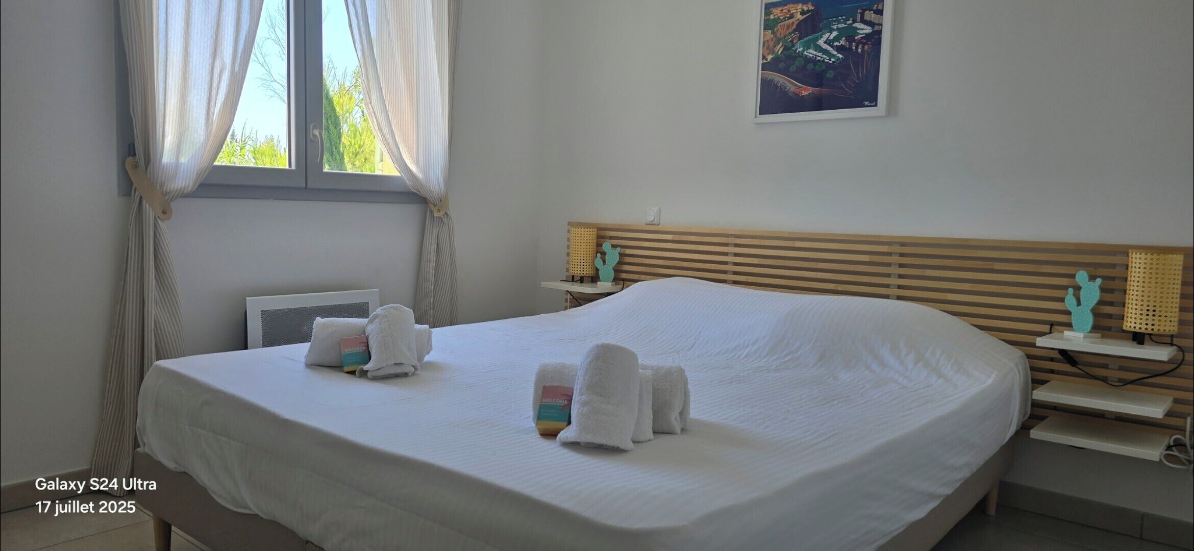A hotel room with a large bed, a bedside table, a nightstand, a window with curtains, and a wall-mounted artwork.