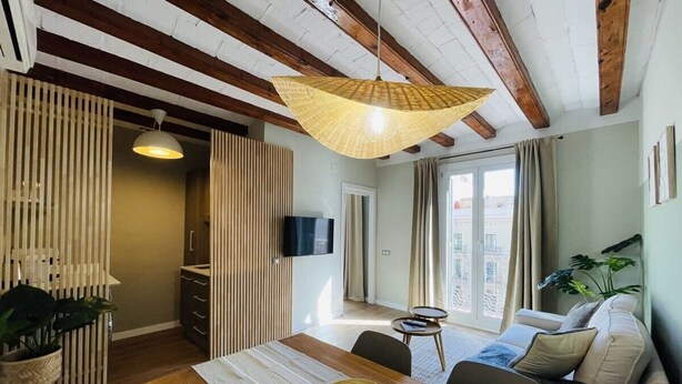 Ein modernes Wohnzimmer mit einem Holzesstisch, einer Couch, einem an der Wand montierten Fernseher und einem großen Fenster mit Vorhängen.
