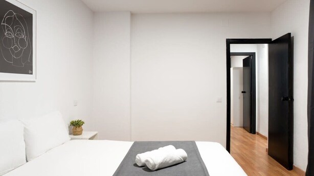 Ein modernes Hotelzimmer mit einem ordentlich bezogenen Bett, einem kleinen Nachttisch mit Pflanze und einer Tür zu einem weiteren Raum.