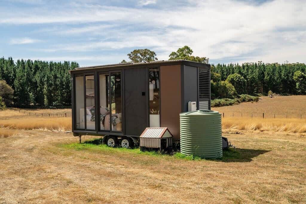 En kompakt, moderne camper med skydedør, et lille tilhørende skur og en vandtank i et landligt område.