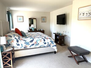 Een slaapkamer met een bed, nachtkastje, bankje, kaptafel, televisie en ingelijste foto's aan de muur.