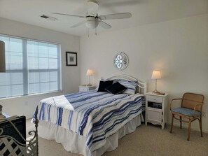 A bedroom with a bed, two bedside tables with lamps, a ceiling fan, a window with blinds, and a decorative wall piece. - Waterview 1st floor 2 bedroom condo with pool access (Ocean View)