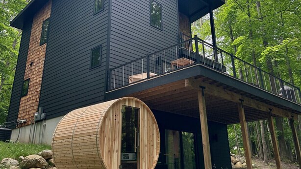 Una cabaña moderna de dos pisos con fachada de madera, un balcón y una gran estructura cilíndrica de madera al frente.