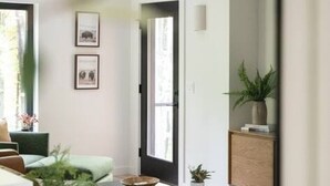 Una sala de estar moderna con un sofá, una mesa de centro y una mesita auxiliar con una planta. Se ve una puerta que da a otra habitación y un mueble bajo con una planta encima.