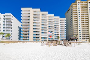 A beachfront hotel with multiple balconies, a sandy beach in front, and a wooden walkway leading to the entrance.