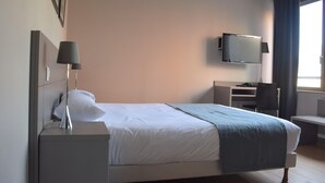 Ein Hotelzimmer mit Bett, Nachttischlampe, Schreibtisch, Stuhl und einem an der Wand befestigten Fernseher.