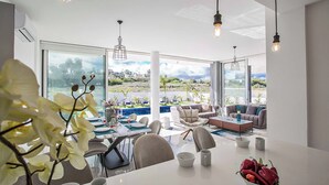 A modern dining area with a view of a pool and landscape, featuring a white table, chairs, and a bowl of fruit.