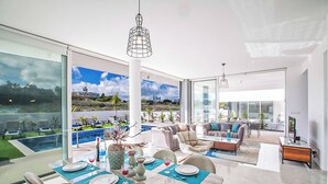 A modern dining area with a table set for four, large windows offering a view of a pool and greenery, and a cozy seating area with a sofa and coffee table.