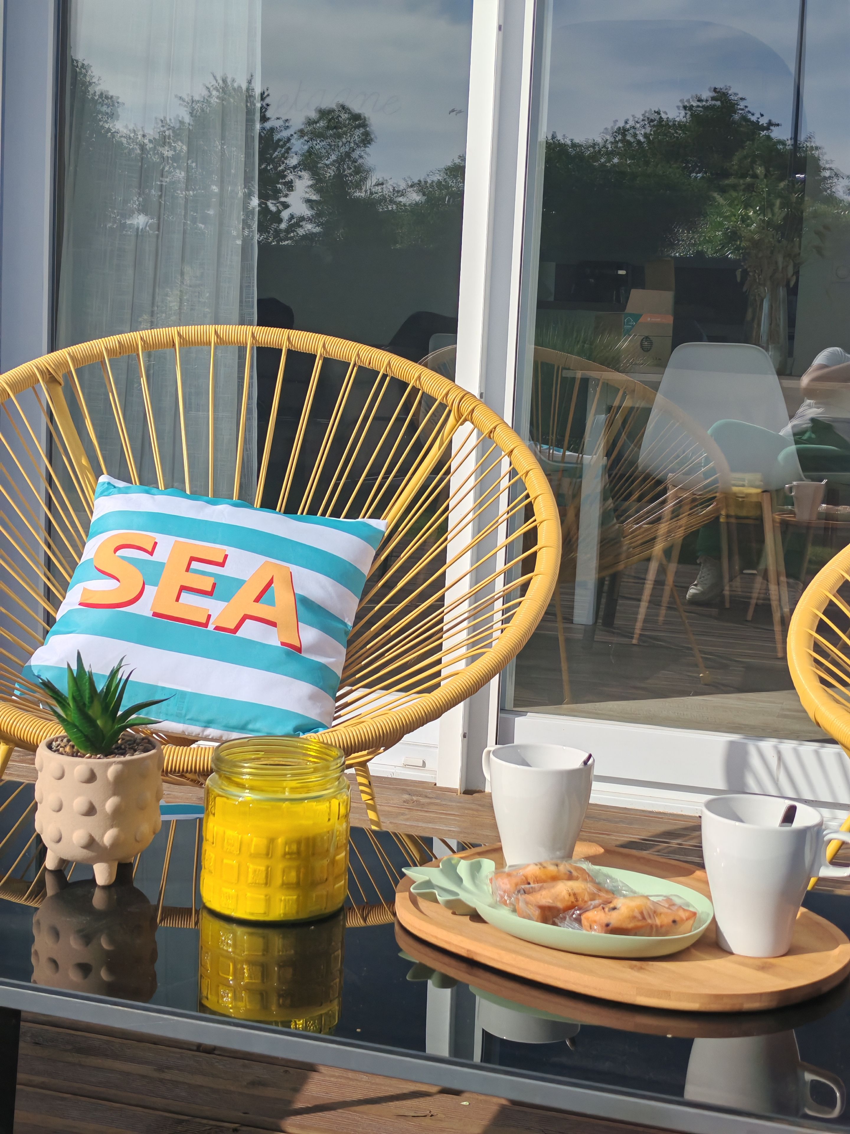 A wicker chair with a cushion labeled 'SEA,' a small potted plant, a jar of yellow liquid, and a tray with a plate of food and cups.