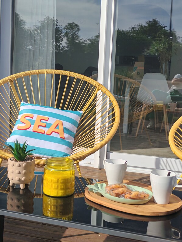 A wicker chair with a cushion labeled 'SEA,' a small potted plant, a jar of yellow liquid, and a tray with a plate of food and cups.